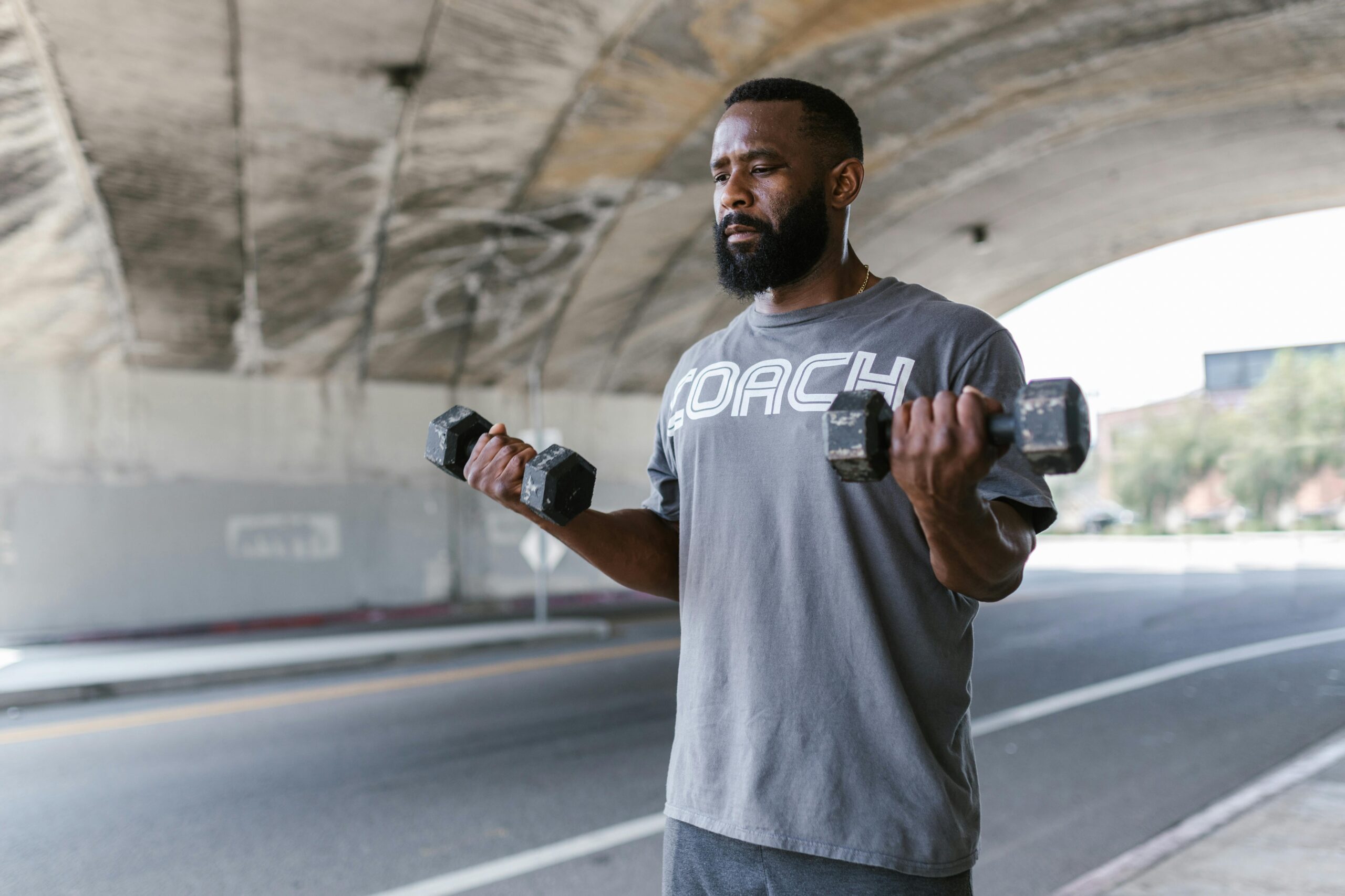 Man lifting dumbbells outdoors under a bridge, emphasizing fitness and strength.