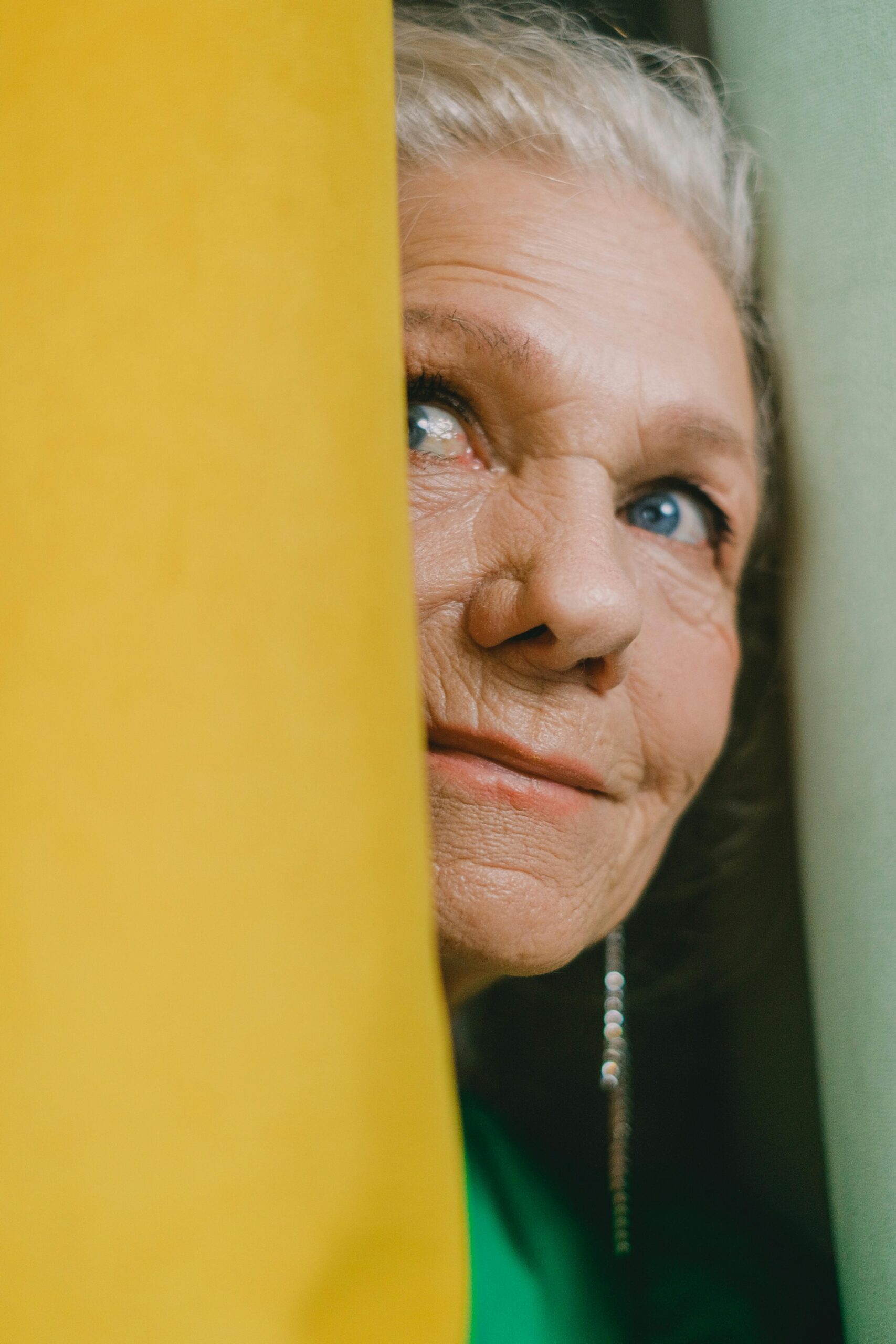 Senior woman smiling gently while peeking through yellow and green curtains, showcasing a thoughtful expression.