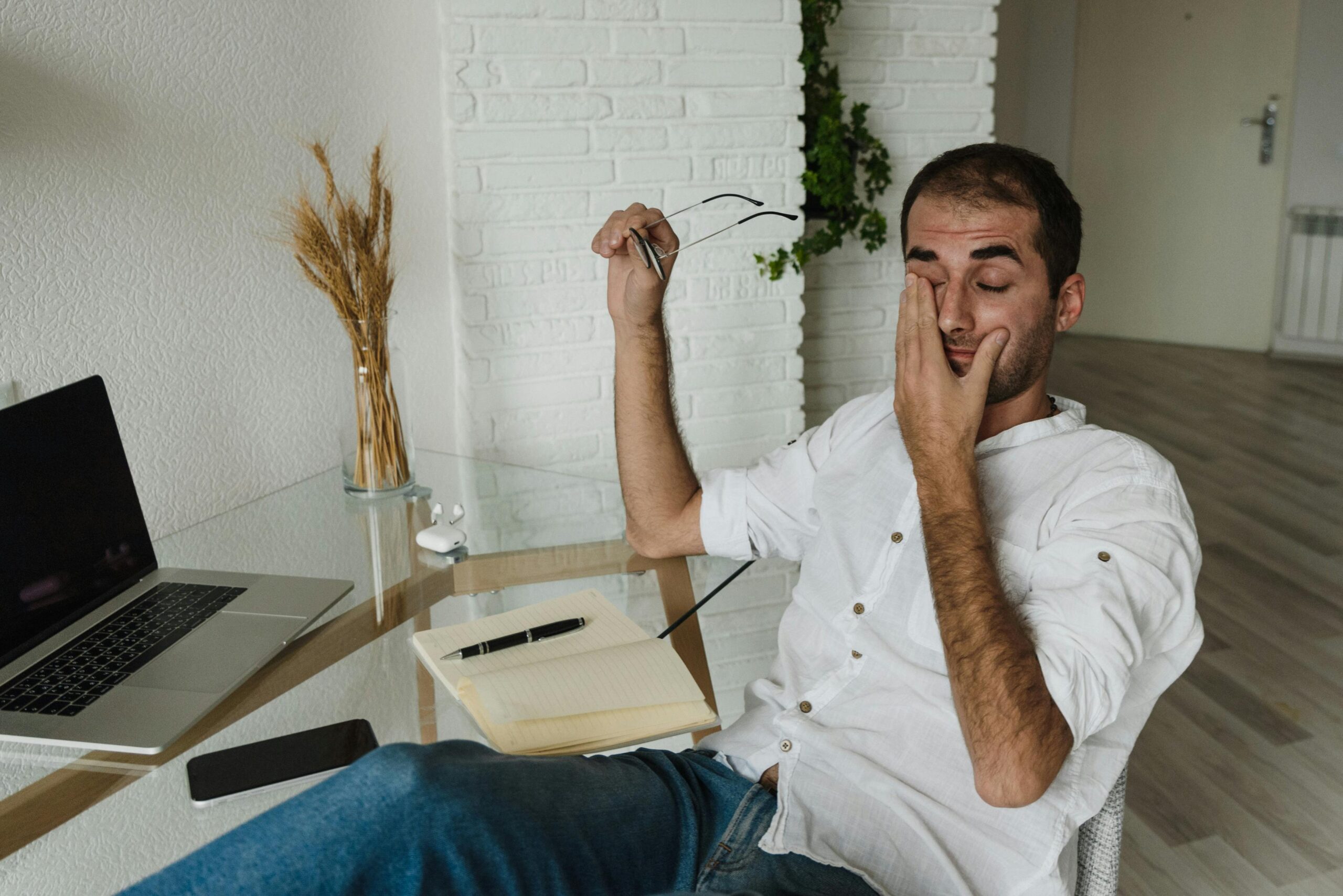 A tired man in a white shirt rests at a home office desk, holding eyeglasses and rubbing his eyes.