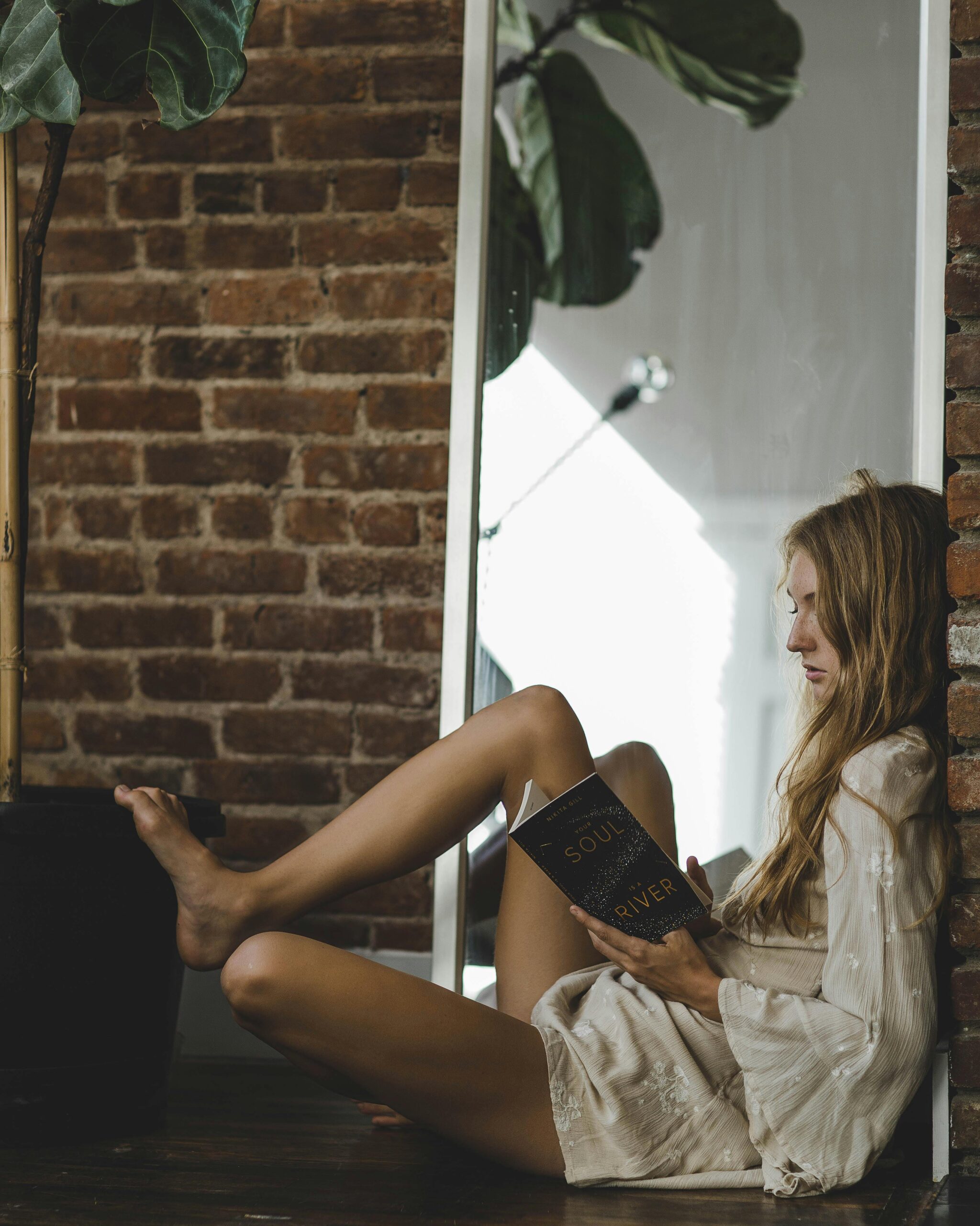 Serene woman reading by a mirror with brick wall backdrop indoors.