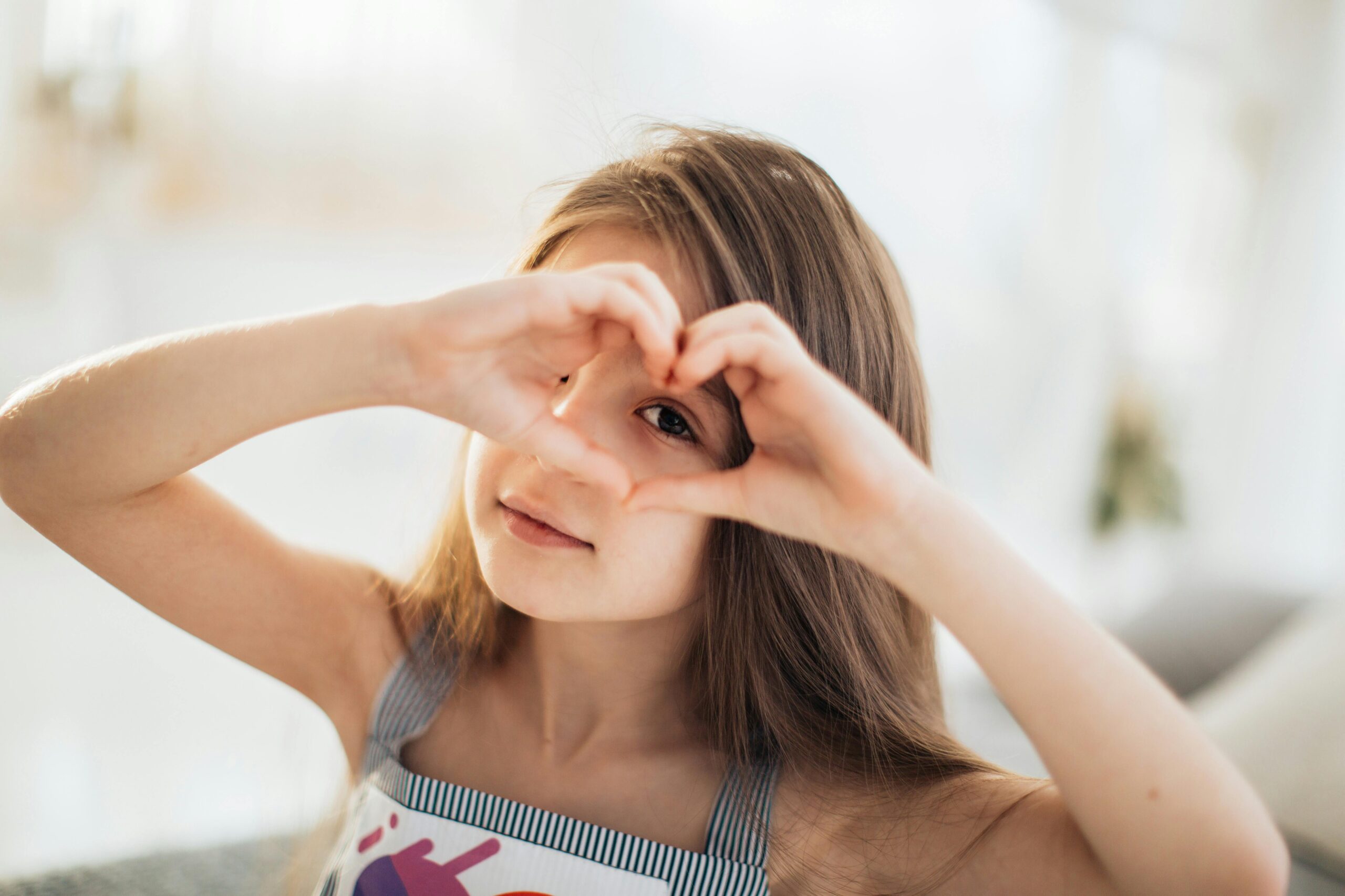 A cute child creating a heart shape with hands indoors, expressing love and warmth.