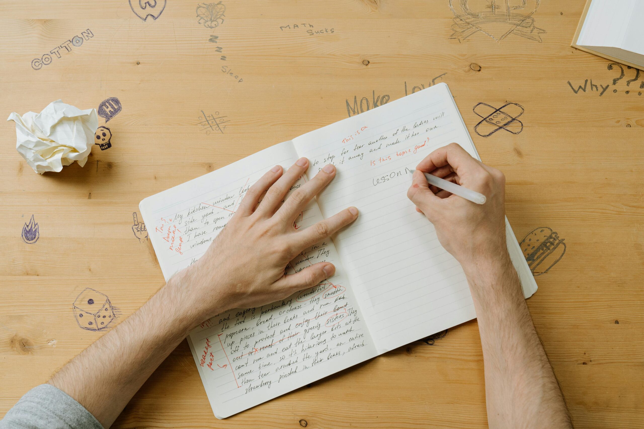 A person writing notes on a notebook with doodles on a wooden desk, reflecting creativity and education.